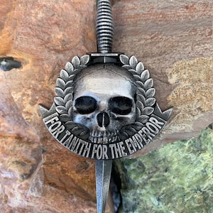 Puede incluir: Una calavera de plata con una corona de laurel alrededor, con una daga que atraviesa la calavera. La daga tiene un mango de plata y una punta afilada. La calavera tiene un diseño detallado y las palabras "For Tanith For The Emperor" están inscritas alrededor de la calavera.