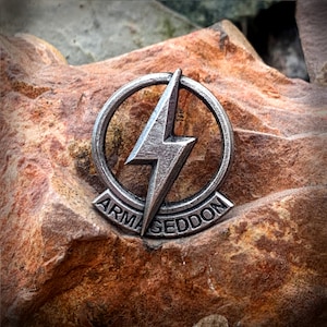Puede incluir: Un pin metálico plateado con un diseño de rayo dentro de un marco circular. La palabra "ARMAGEDDON" está inscrita en una pancarta debajo del rayo. El pin está sobre un fondo de piedra marrón texturizada.