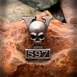 May include: Silver skull pendant with a V-shaped design and the number 597 on a rectangular plaque.