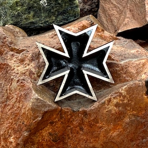 Puede incluir: Emblema de la Cruz de Hierro negro y plateado. La cruz tiene un centro negro con un borde plateado y está colocada sobre un fondo de rocas marrones y verdes. El emblema es una cruz de cuatro puntas con brazos acampanados.