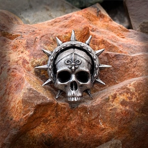 May include: A silver skull with a fleur-de-lis on its forehead is surrounded by a spiked halo. The skull is on a rough, brown, textured surface.