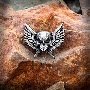 May include: A silver skull with wings pin. The skull has a detailed design with hollow eye sockets and a prominent jawline. The wings are spread out behind the skull and have a textured surface.