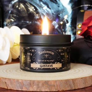 May include: A lit black candle in a tin with the name "Gustave" on a wooden slice. The candle has a label that reads "Nookworm" and "Get out of my way!" with a weight of 4 oz (110 g).