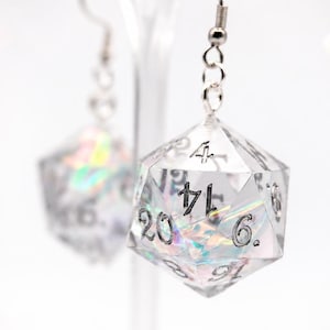 May include: A pair of dangle earrings featuring clear, twenty-sided polyhedral dice with iridescent, holographic-like inclusions inside. Each die has black engraved numbers and is attached to a silver-toned metal fishhook ear wire.