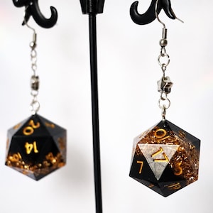 May include: A pair of dangle earrings featuring 20-sided dice as pendants. The dice are black with gold-colored flakes embedded inside and gold-colored numbers. Each die hangs from a silver-toned metal chain with a small star-shaped charm and hook.
