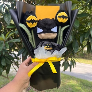 May include: A bouquet featuring a central crocheted Batman figure flanked by two crocheted tulips with yellow centers and black bat symbols. The arrangement is wrapped in black paper with white tissue paper and tied with a bright yellow ribbon.