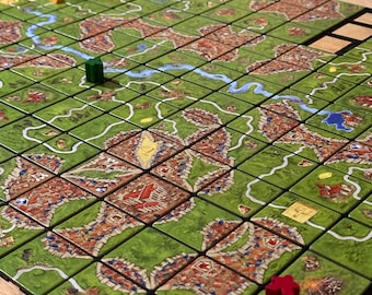 May include: A board game in progress, featuring green tiles with roads, cities, and fields. Wooden game pieces in red, blue, yellow, and green are placed on the board. The game is set on a wooden table.