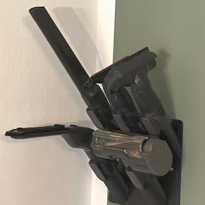 Può includere: Un supporto nero per accessori per aspirapolvere montato a parete. Il supporto ha più fessure e clip per riporre vari accessori, tra cui un'asta lunga, una testina e un'unità portatile. Il supporto è progettato per mantenere gli accessori dell'aspirapolvere organizzati e accessibili.