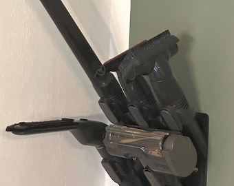 May include: A black vacuum cleaner accessory holder mounted on a wall. The holder has multiple slots and clips to store various attachments, including a long wand, a brush head, and a handheld unit. The holder is designed to keep vacuum accessories organized and accessible.