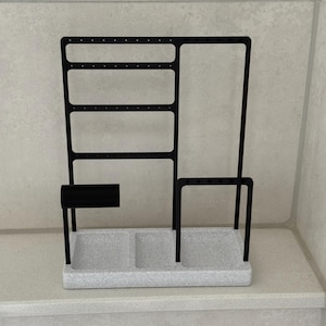 May include: Black metal jewelry organizer with a speckled gray base. The organizer features a section for earrings, a bar, and a tray with two compartments. The design is modern and minimalist.