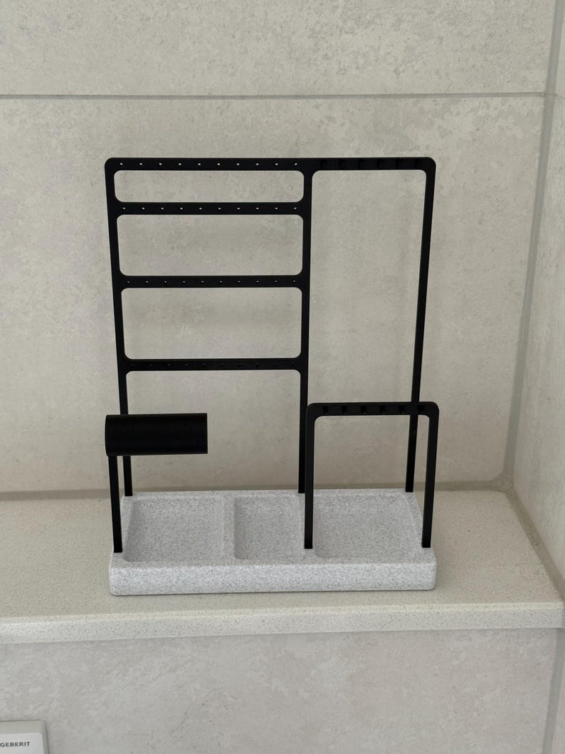 May include: Black metal jewelry organizer with a speckled gray base. The organizer features a section for earrings, a bar, and a tray with two compartments. The design is modern and minimalist.