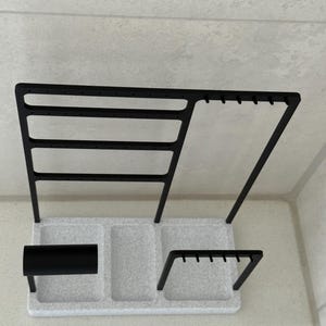 May include: A black metal jewelry organizer with a speckled white base. The organizer has multiple sections for storing earrings, necklaces, and other accessories. The design includes a bar for bracelets and a section for rings.