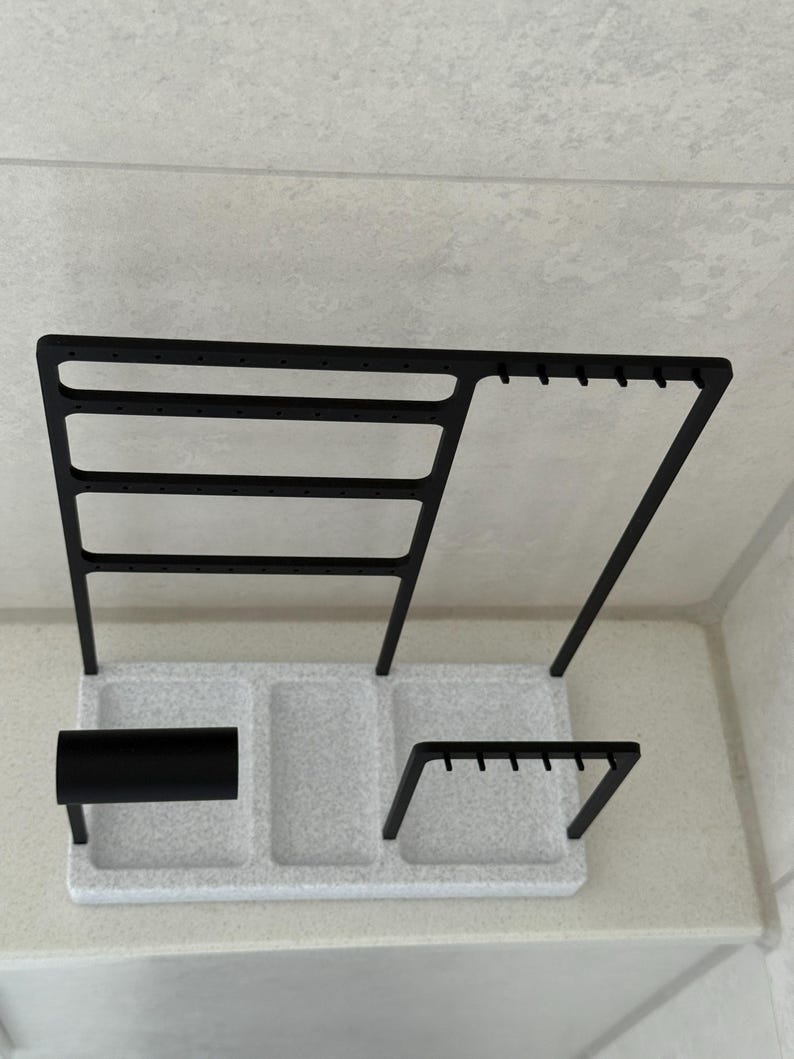 May include: A black metal jewelry organizer with a speckled white base. The organizer has multiple sections for storing earrings, necklaces, and other accessories. The design includes a bar for bracelets and a section for rings.