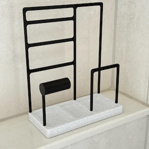 May include: A black metal jewelry organizer with a speckled white base. Features include earring slots, a bracelet holder, and two vertical bars. The organizer is designed to hold various types of jewelry.