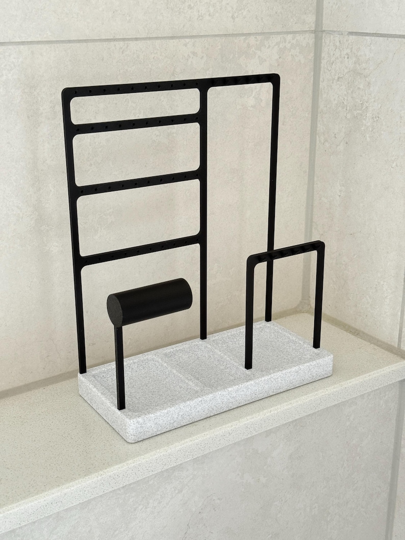 May include: A black metal jewelry organizer with a speckled white base. Features include earring slots, a bracelet holder, and two vertical bars. The organizer is designed to hold various types of jewelry.