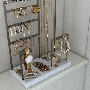 May include: A gray metal jewelry organizer filled with gold necklaces, earrings, bracelets, and a watch. The organizer has multiple levels and compartments to display various jewelry pieces. A pearl hair clip and a gold chain are also visible.