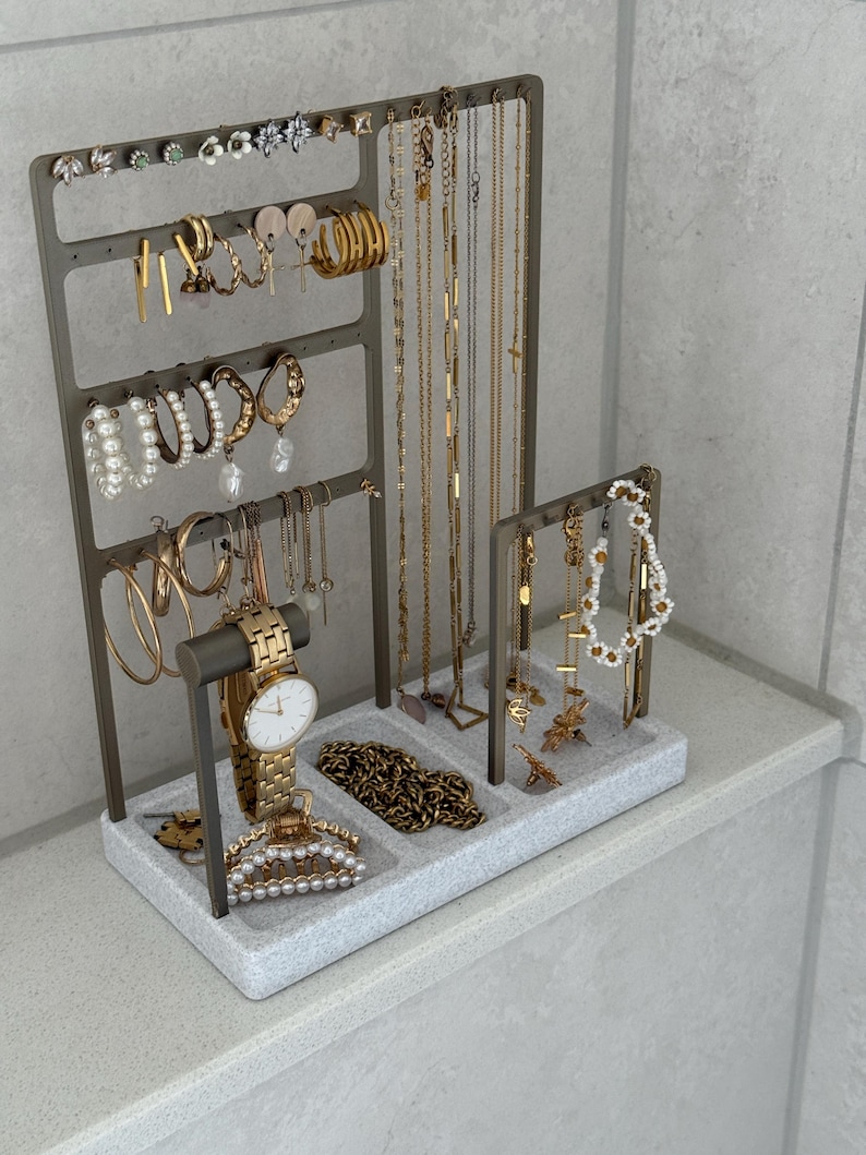 May include: A gray metal jewelry organizer filled with gold necklaces, earrings, bracelets, and a watch. The organizer has multiple levels and compartments to display various jewelry pieces. A pearl hair clip and a gold chain are also visible.