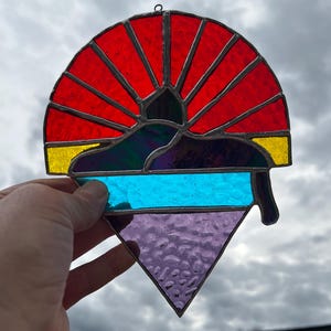 Könnte beinhalten: Ein Glasfenster-Sonnenfänger mit einem roten, gelben, blauen, lila und grünen Design. Der Sonnenfänger hat eine halbkreisförmige Oberseite mit roten Strahlen, einen blauen horizontalen Streifen und ein lila Dreieck am Boden. Er wird vor einem bewölkten Himmel gehalten.