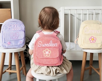 May include: Three small, personalized backpacks in purple, pink, and yellow. The backpacks have embroidered flowers and text. The text on the purple backpack says "Elle", the text on the pink backpack says "Sweden", and the text on the yellow backpack says "Emma".