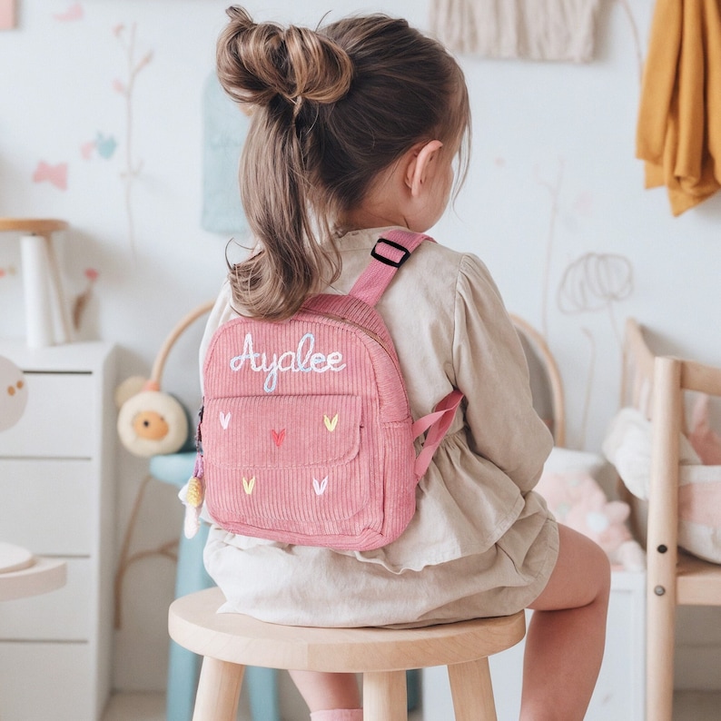 May include: A pink corduroy backpack with the name "Ayalee" embroidered on the front. The backpack has a small pocket on the front and a zipper closure. The backpack is being worn by a young girl with long brown hair.