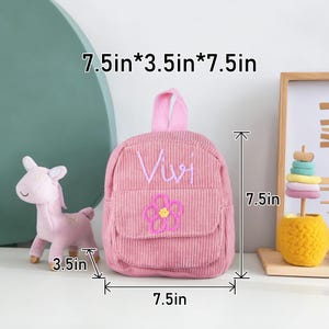 May include: Pink corduroy mini backpack with a flower and the name "Vivi" embroidered on the front. The backpack measures 7.5 inches wide, 3.5 inches deep, and 7.5 inches tall.