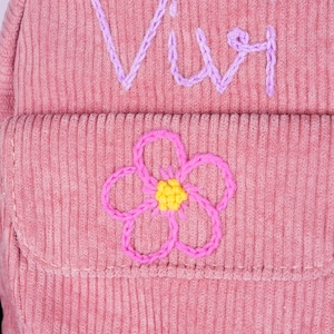 May include: Pink corduroy backpack with a flower embroidered on the front pocket and the word "Viv" embroidered on the top flap.