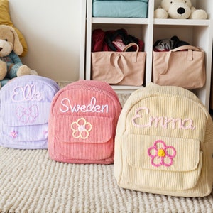 K&ouml;nnte beinhalten: Drei Cord-Rucks&auml;cke mit gestickten Namen und Blumen. Die Rucks&auml;cke sind lavendelfarben, rosa und cremefarben.
