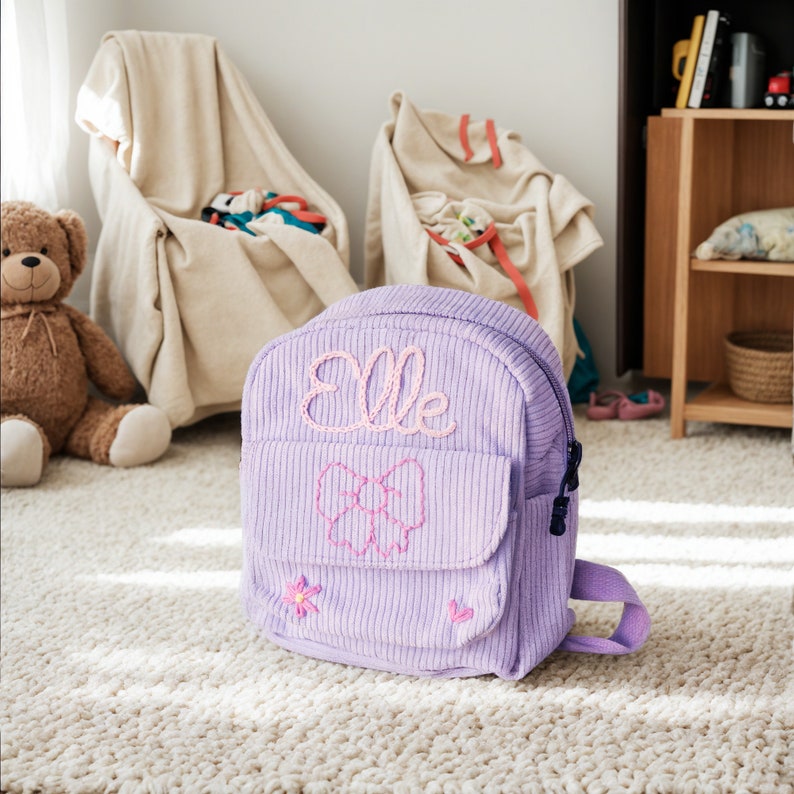 K&ouml;nnte beinhalten: Ein lilafarbener Cordrucksack mit einer rosa gestickten Schleife und dem Namen "Elle" auf der Vorderseite.
