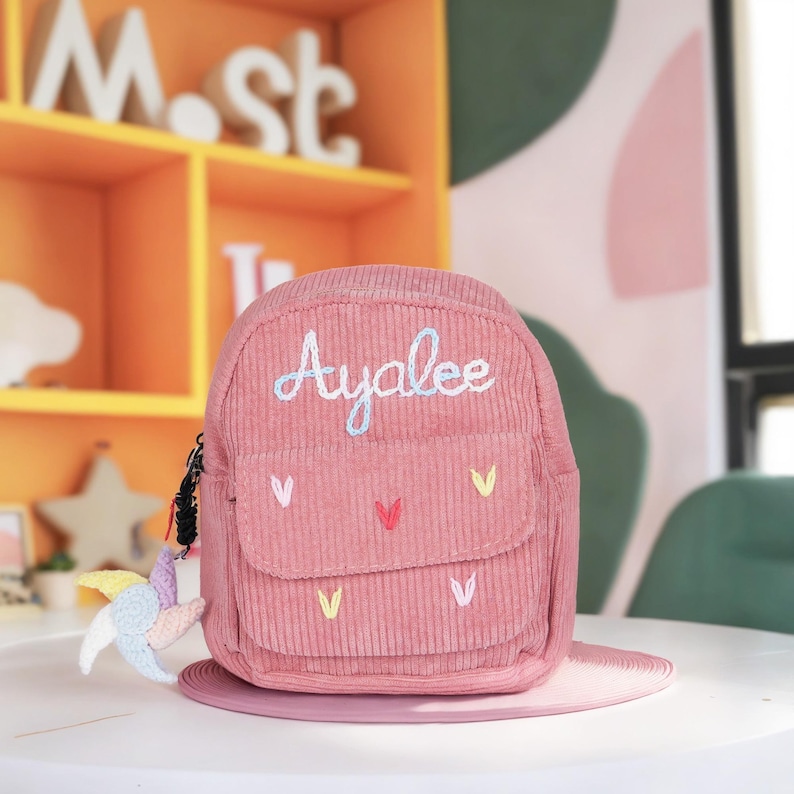 May include: A pink corduroy mini backpack with the name "Ayalee" embroidered on the front. The backpack has a small pocket on the front and a zipper closure. There are small embroidered hearts in various colors on the front of the backpack.