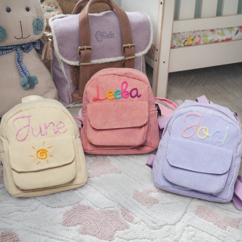 May include: Three small, personalized backpacks in different colors. The backpacks are made of corduroy and have embroidered names on the front. The names are June, Leeba, and Jodi.