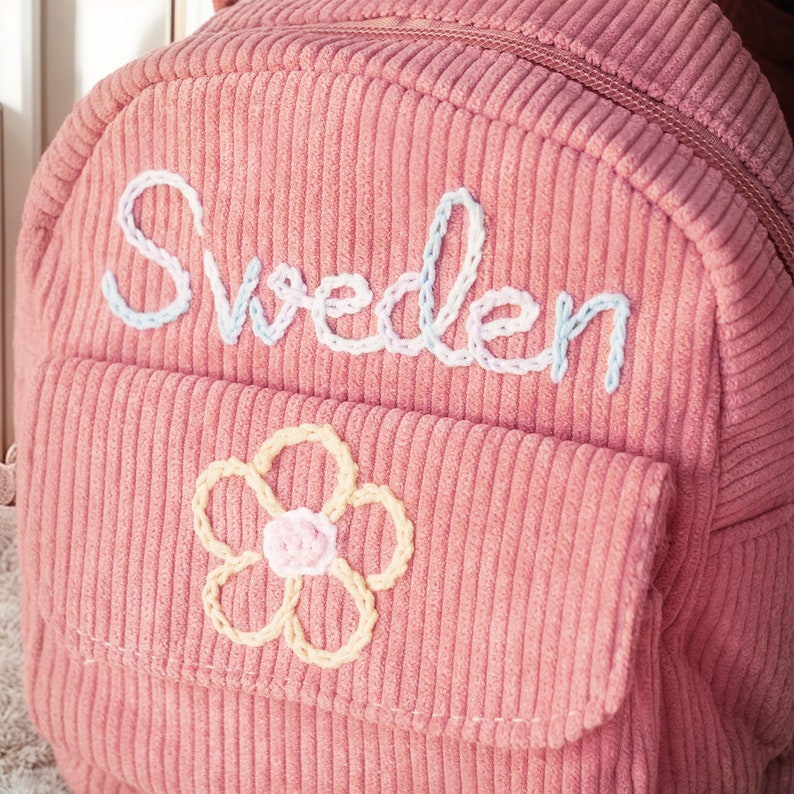 K&ouml;nnte beinhalten: Ein rosa Cordrucksack mit dem Wort "Schweden" in wei&szlig;er Stickerei auf der Vorderseite. Eine gelbe Blume mit pinkfarbenem Zentrum ist ebenfalls auf der Vorderseite gestickt.