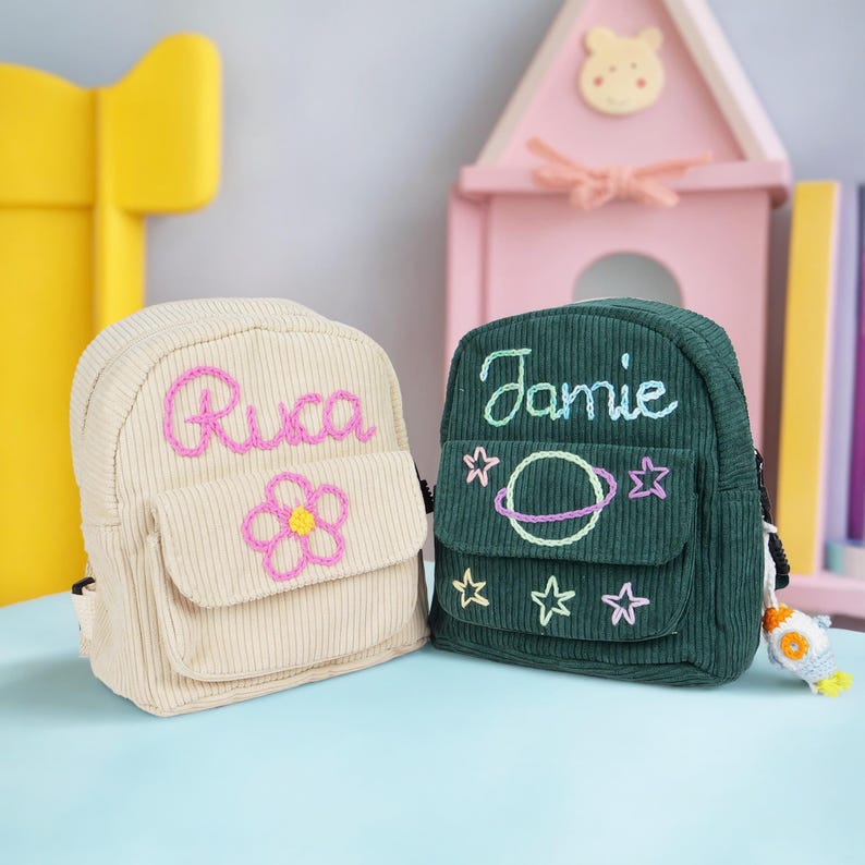 May include: Two small, personalized backpacks. One is beige with the name "Rica" embroidered in pink and a pink flower. The other is green with the name "Jamie" embroidered in white and a planet with stars.