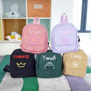 May include: Five small, personalized corduroy backpacks in different colors. The backpacks have embroidered designs, including a flower, a bow, a crown, a car, and another flower. The backpacks are labeled with the names Vivi, Ameera, Chance, Yonah, and Daisy.