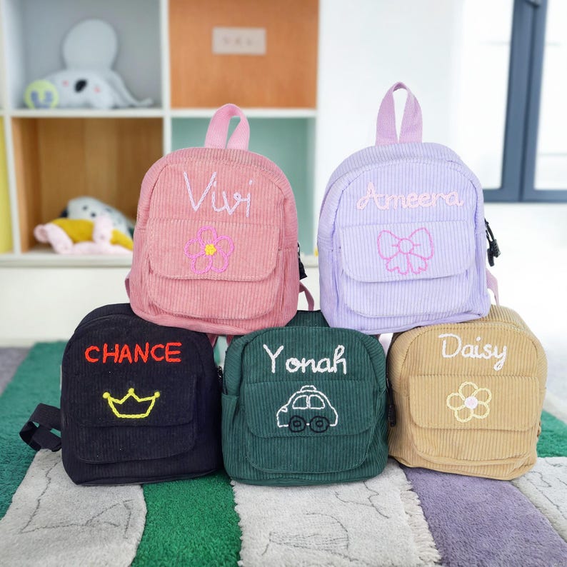 May include: Five small, personalized corduroy backpacks in different colors. The backpacks have embroidered designs, including a flower, a bow, a crown, a car, and another flower. The backpacks are labeled with the names Vivi, Ameera, Chance, Yonah, and Daisy.