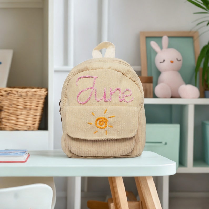 May include: A beige corduroy backpack with a pink embroidered name "June" and a yellow embroidered sun on the front pocket.