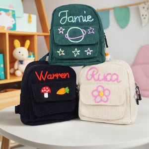 May include: Three small, personalized backpacks in green, black, and white corduroy. The green backpack has a planet and stars embroidered on it and says "Jamie". The black backpack has a mushroom and carrot embroidered on it and says "Warren". The white backpack has a flower embroidered on it and says "Rica".