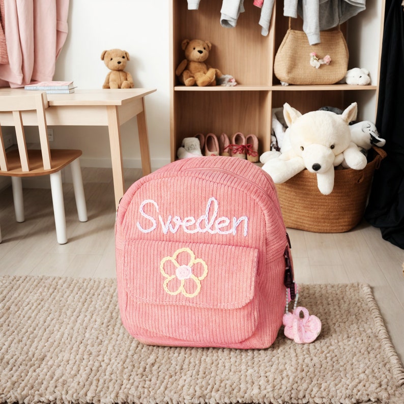 K&ouml;nnte beinhalten: Rosa Cord-Rucksack mit einer wei&szlig;en gestickten Blume und dem Wort "Schweden" auf der Vorderseite.