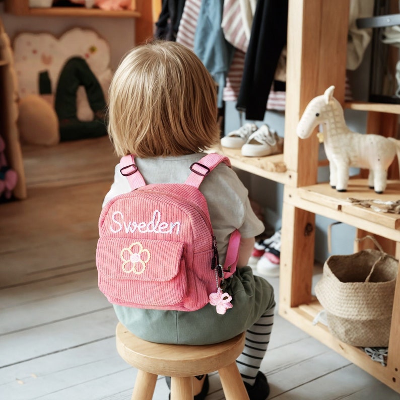 K&ouml;nnte beinhalten: Ein rosa Cordrucksack mit einer wei&szlig;en gestickten Blume und dem Wort "Schweden" auf der Vorderseite. Der Rucksack wird von einem Kind getragen, das auf einem Holzhocker sitzt.