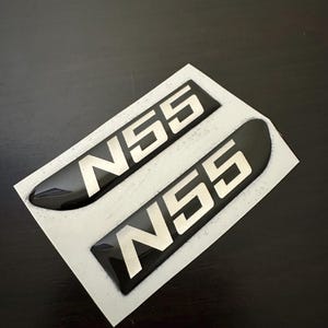 Pode incluir: Dois autocolantes de emblema N55 pretos e prateados sobre um suporte branco. Os autocolantes têm um acabamento brilhante e uma forma retangular com bordas arredondadas. O texto "N55" é prateado e em negrito.