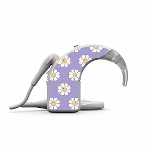 Könnte beinhalten: Ein weißes Hörgerät mit einem lila und weißen Gänseblümchenmuster. Das Gänseblümchenmuster zeigt ein gelbes Smiley-Gesicht in der Mitte jeder Blume.