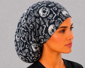 Op de afbeelding: Een zwarte operatiemuts met een herhalende witte grafische print van menselijke schedels, skeletachtige handen en oogbollen. De stoffen hoofdbedekking heeft een afgeronde, volumineuze vorm met een gladde band aan de voorzijde.