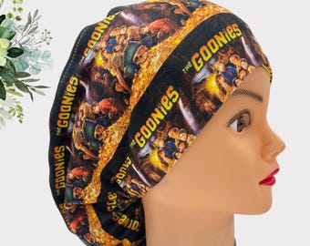 May include: A black scrub hat with a colorful print of the Goonies movie characters. The hat has a yellow and black striped pattern with the words "The Goonies" repeated throughout.
