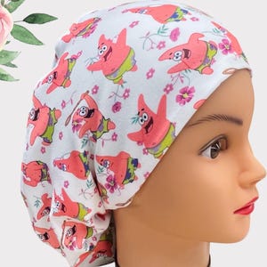 May include: White scrub hat with a pink and white pattern of cartoon characters. The characters are wearing pink and green clothing and have a pink star on their chest. The hat is made of a soft, comfortable fabric.
