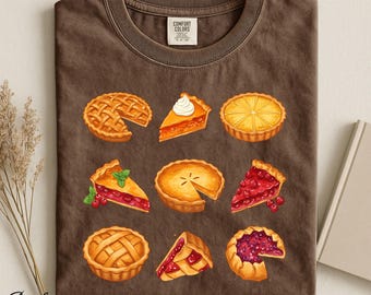 Op de afbeelding: Een bruin t-shirt met ronde hals en een grafische print van negen verschillende taarten in een raster. De illustraties tonen diverse fruit- en custardtaarten met rasterkorsten, punten en hele taarten in tinten rood, oranje en geel.