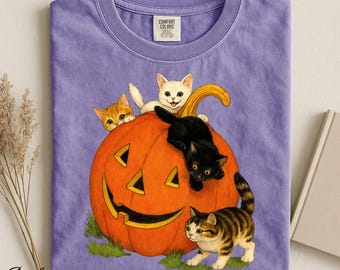 Op de afbeelding: Een paars t-shirt met een afbeelding van een grote oranje pompoen met een uitgesneden gezicht, omringd door vier kittens. Eén wit kitten zit bovenop, een zwart kitten klimt aan de zijkant en twee cyperse kittens staan aan de onderkant.