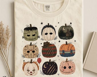 Op de afbeelding: Een ivoorkleurig t-shirt met een grafische print van negen gestileerde pompoenen die zijn versierd als diverse horrorfilmfiguren en thema's, waaronder een monster met stiksels, een ijshockeymasker, een spookhuis en een clowngezicht.