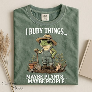 Może przedstawiać: Koszulka w kolorze mchu z grafiką żaby w słomkowym kapeluszu i ogrodniczkach, trzymającej łopatę. Tekst brzmi I BURY THINGS... MAYBE PLANTS... MAYBE PEOPLE. Wz&oacute;r zawiera rośliny doniczkowe oraz dłoń wyłaniającą się z ziemi na dole grafiki.