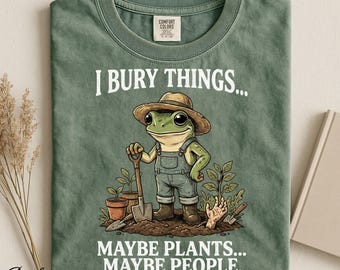 Op de afbeelding: Een mosgroen t-shirt met een afbeelding van een kikker in een strooien hoed en tuinbroek, die een schep vasthoudt. De tekst luidt I BURY THINGS... MAYBE PLANTS... MAYBE PEOPLE. Het ontwerp bevat potplanten en een hand die uit de grond komt.