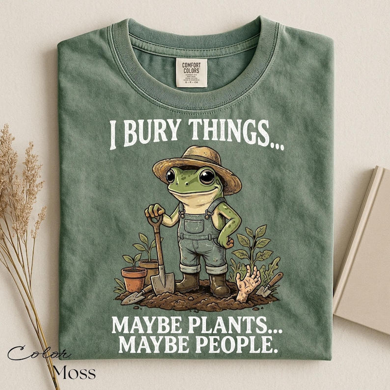 Może przedstawiać: Koszulka w kolorze mchu z grafiką żaby w słomkowym kapeluszu i ogrodniczkach, trzymającej łopatę. Tekst brzmi I BURY THINGS... MAYBE PLANTS... MAYBE PEOPLE. Wz&oacute;r zawiera rośliny doniczkowe oraz dłoń wyłaniającą się z ziemi na dole grafiki.