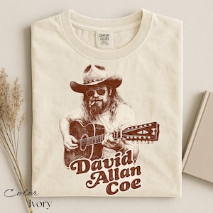 May include: Ivory-colored t-shirt featuring a sepia-toned illustration of a musician in a cowboy hat playing a guitar. The text "David Allan Coe" is printed in a retro font below the image. The shirt has a crew neck and a Comfort Colors tag.